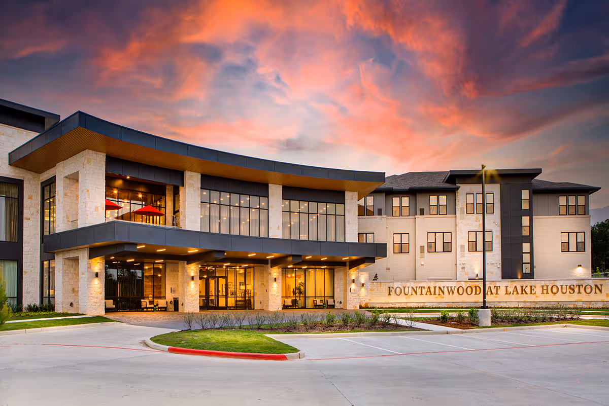 Exterior view of Fountainwood at Lake Houston building during sunset with a dramatic pink and purple sky. The modern building features large windows, stone and dark paneling, and a well-lit entrance with outdoor seating. A sign with the facility name is visible on the right side.