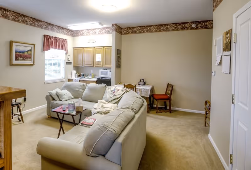 A cozy living room area with a light gray sectional sofa, a small coffee table with a tissue box and a glass, and a small dining table with two chairs against a beige wall. The room has beige carpet, a window with blinds and a red valance, a framed landscape painting on the wall, and a small kitchen area with wooden cabinets and appliances in the background.