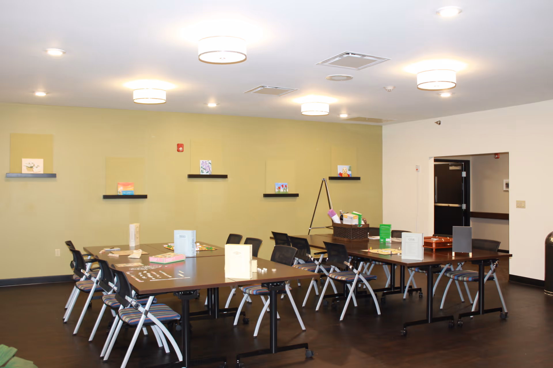 Spacious activity/dining room with tables and chairs, small artworks on wall shelves, and overhead lighting.