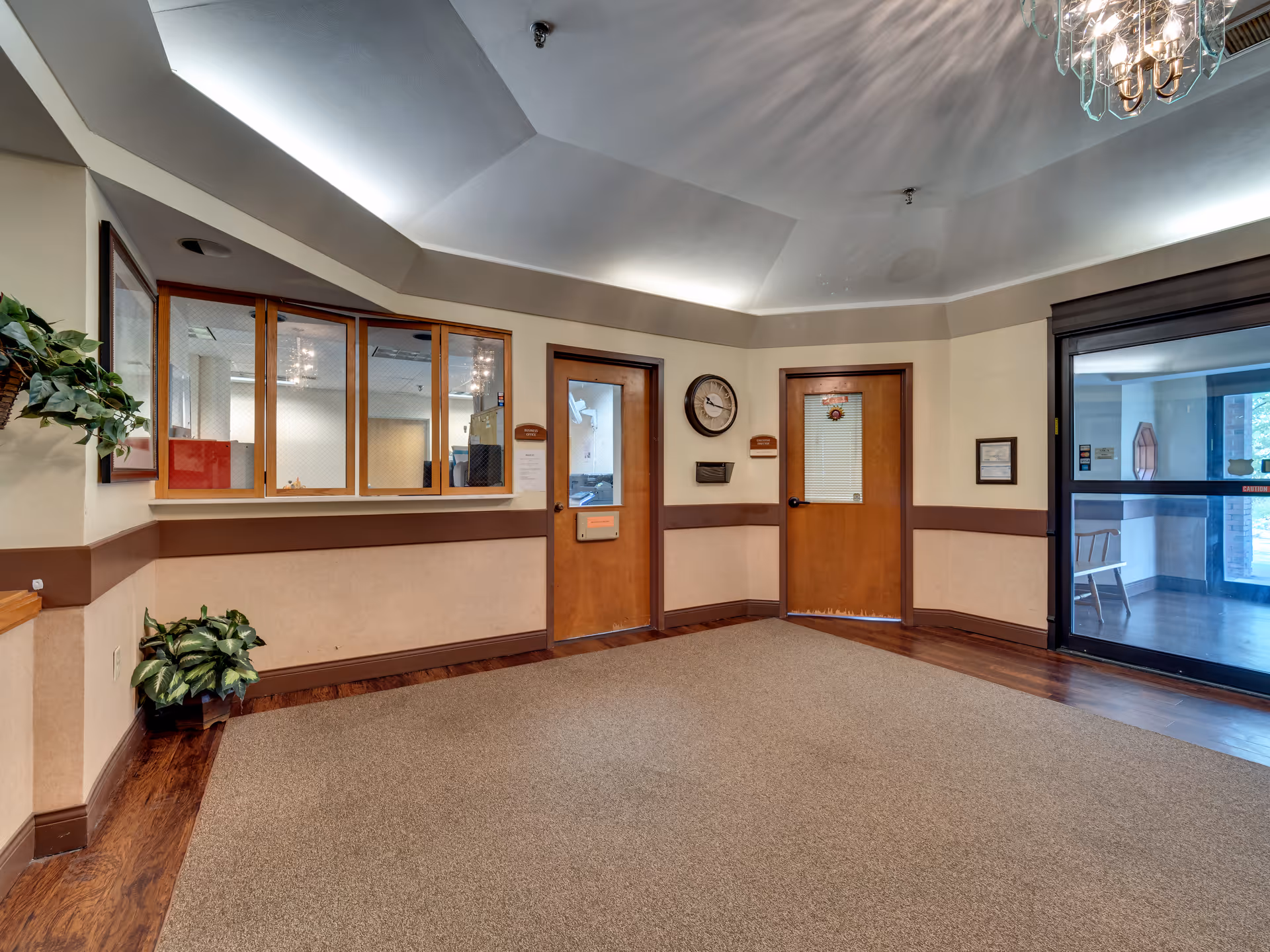 Reception lobby with service windows, two wooden doors, a wall clock, potted plants, and a glass entry door.
