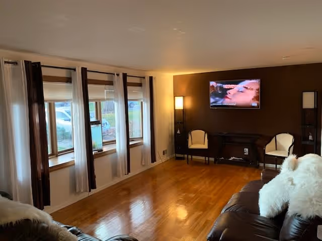 A spacious living room with polished wooden floors, a large window with white and dark brown curtains, a wall-mounted TV showing a close-up scene, two chairs with cushions, a small table, and a dark brown leather sofa with white fluffy pillows.