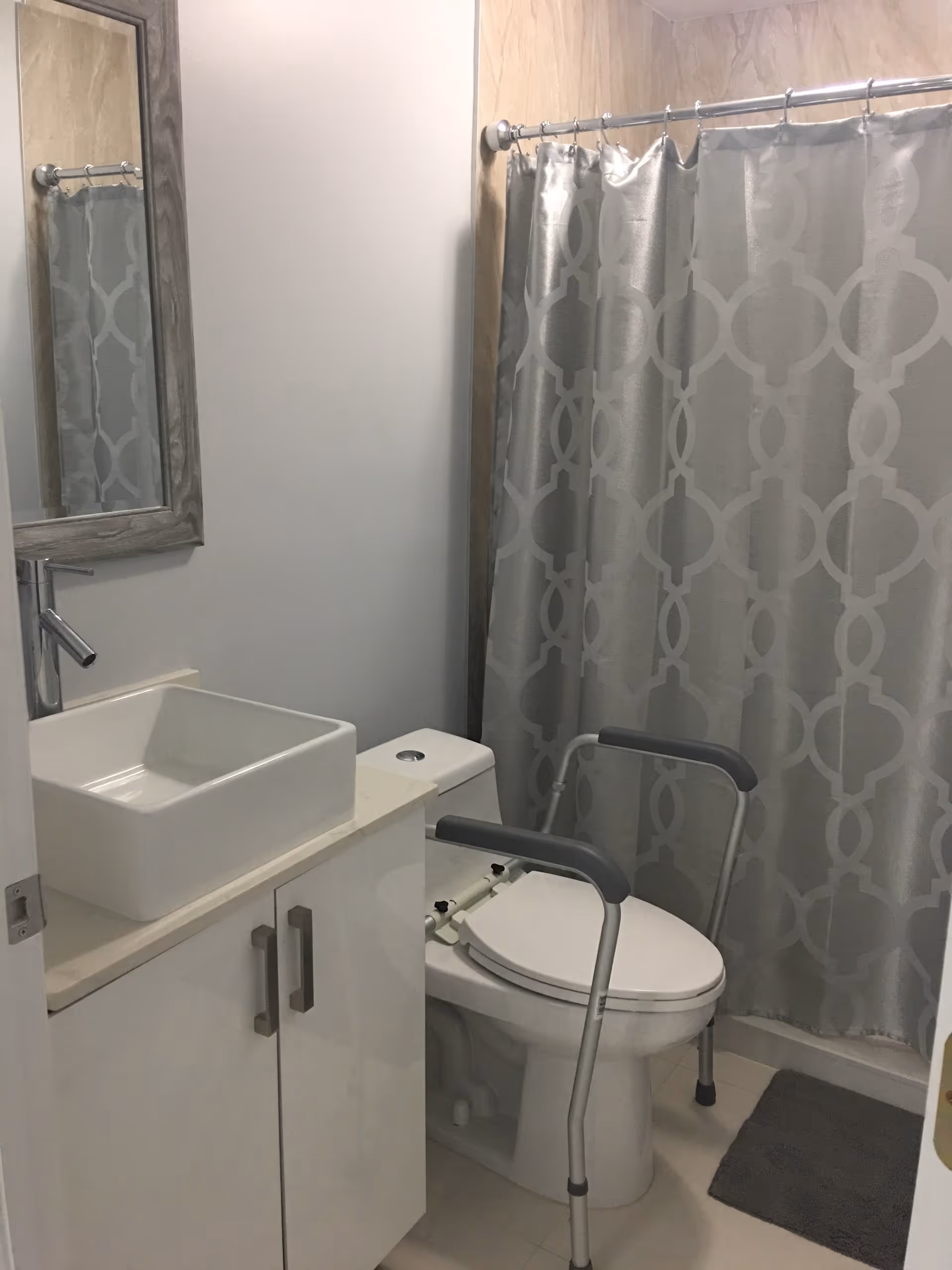 A small bathroom with a white sink on a cabinet, a toilet with a gray safety frame around it, a gray patterned shower curtain, and a gray bath mat on the floor.