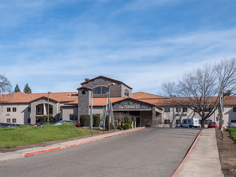 Exterior view of a senior living facility named The Terraces, showing a large building with a red-tiled roof, a driveway leading up to the entrance, several parked cars, and a clear blue sky.