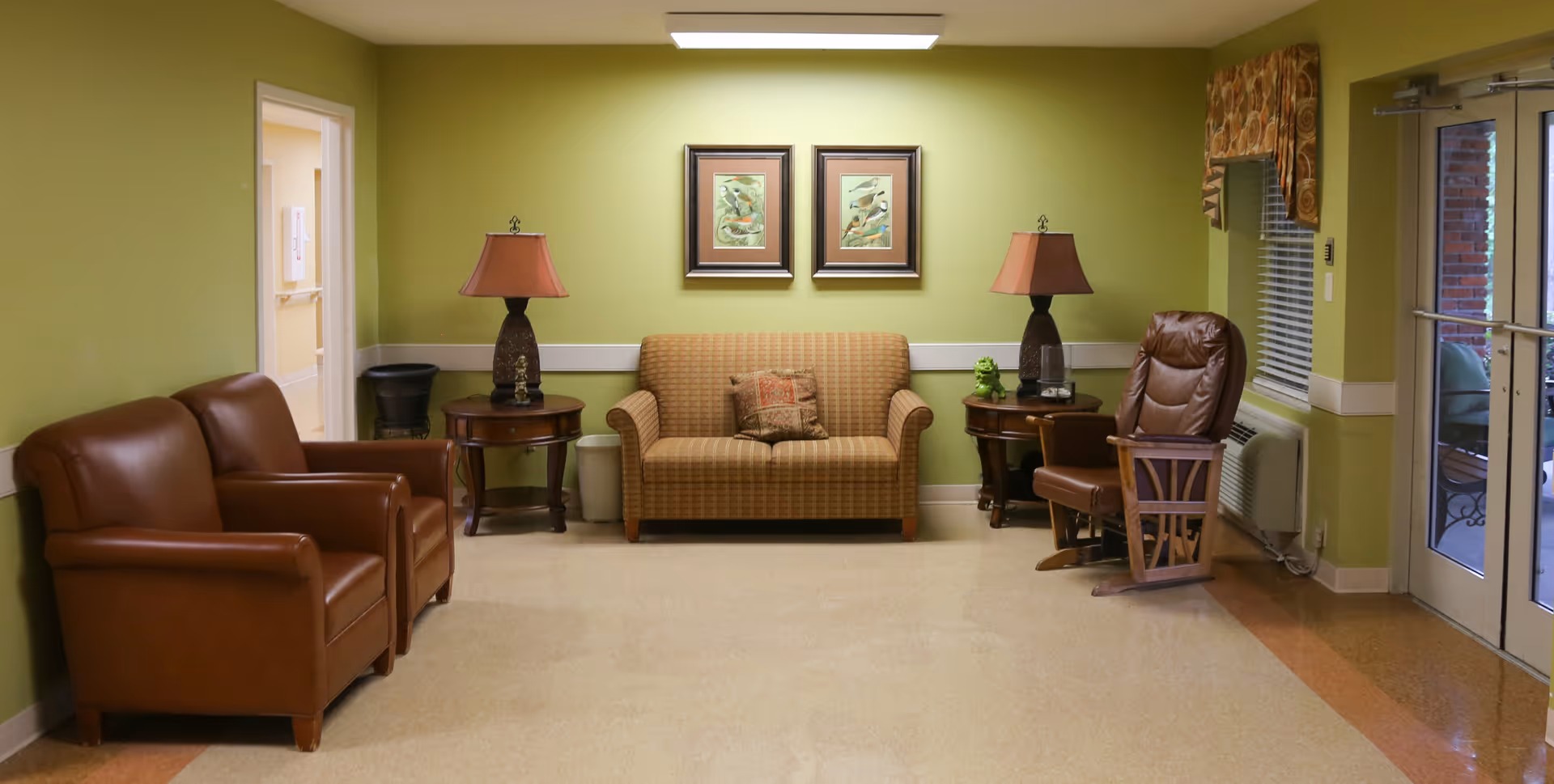 Small common seating area with leather chairs, a loveseat, side tables and lamps against a green wall near glass doors.