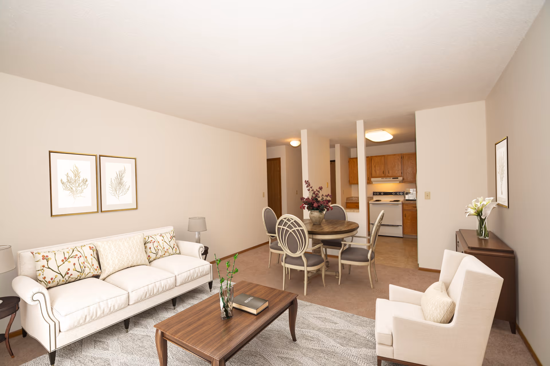 A cozy living room and dining area in a senior living facility with a white sofa adorned with patterned cushions, a wooden coffee table with a book and a small plant, a round dining table with four chairs and a flower vase centerpiece, and a small kitchen with wooden cabinets and white appliances in the background. The walls are beige with framed botanical prints, and there is a beige armchair with a cushion near a wooden sideboard with a vase of white flowers.