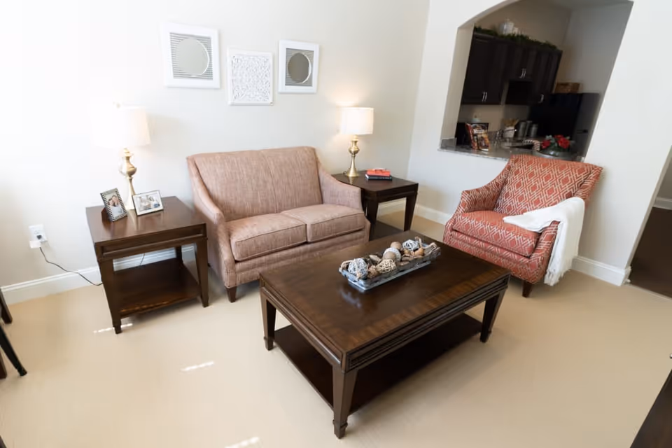 A cozy living room area with a brown loveseat, a patterned red armchair with a white throw draped over one arm, two wooden side tables each with a lamp, and a wooden coffee table with decorative items. The room has light-colored walls and carpet, with three framed wall decorations above the loveseat. A kitchen area is visible through an open pass-through window.