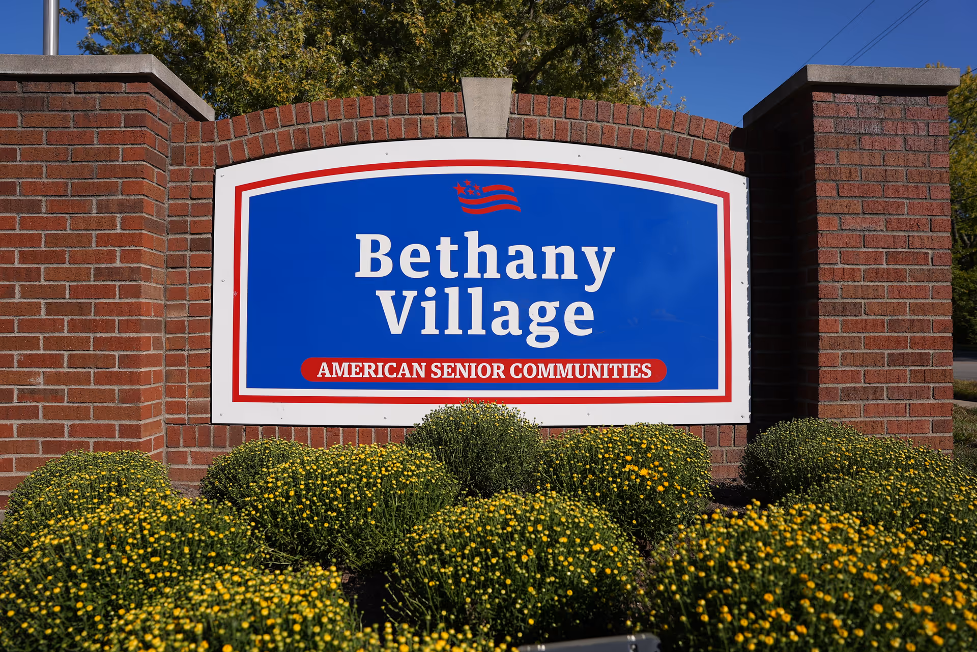 A large blue and white sign with red accents reading 'Bethany Village AMERICAN SENIOR COMMUNITIES' mounted on a brick structure with green bushes and yellow flowers in front, under a clear blue sky.
