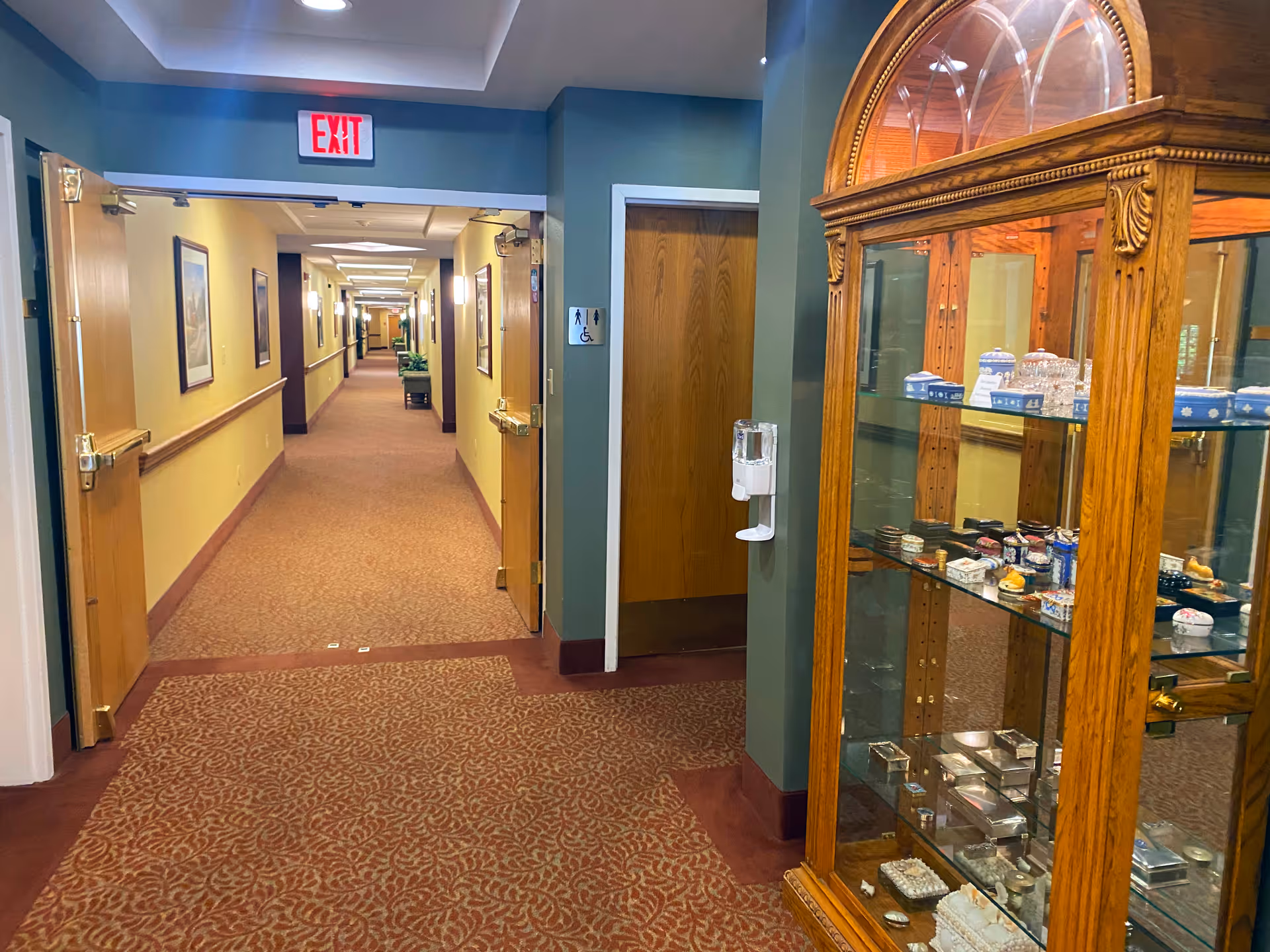 A long, carpeted hallway in a senior living facility with yellow walls and wooden doors on both sides. There is an exit sign above an open doorway at the near end of the hallway. On the right side, there is a wooden display cabinet with glass shelves holding various decorative items. A hand sanitizer dispenser is mounted on the wall near a restroom door marked with accessibility signs.