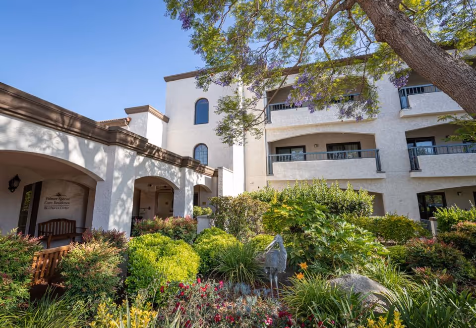 Exterior view of Casa de las Campanas facility showing a white multi-story building with balconies, surrounded by lush green bushes, flowering plants, and a tree with purple flowers under a clear blue sky.