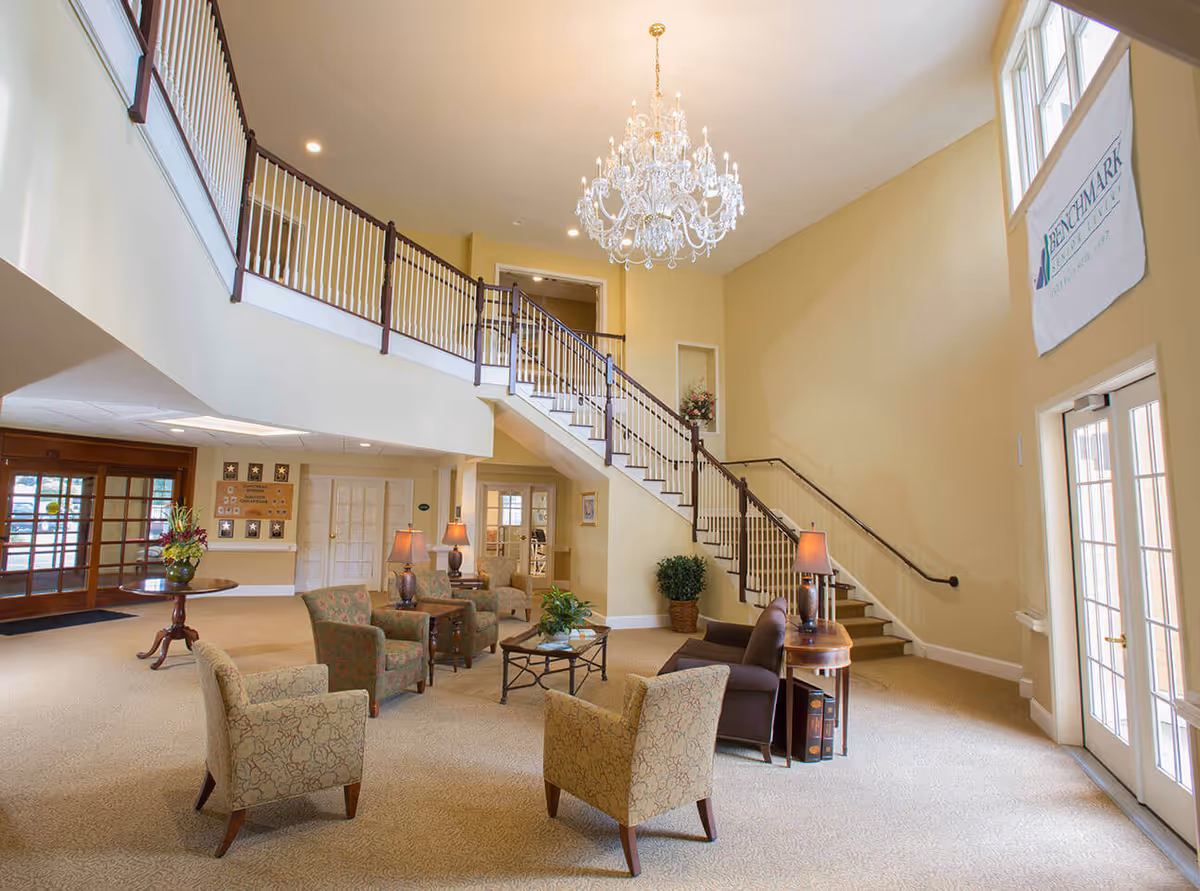 A spacious and well-lit senior living facility lobby with beige walls and carpet. The area features a large crystal chandelier hanging from the ceiling, a staircase with white railings and dark wood handrails leading to an upper floor, and several upholstered chairs arranged around small tables with lamps and plants. There is a large window and glass doors letting in natural light, and a banner on the wall that reads 'Benchmark Senior Living'.