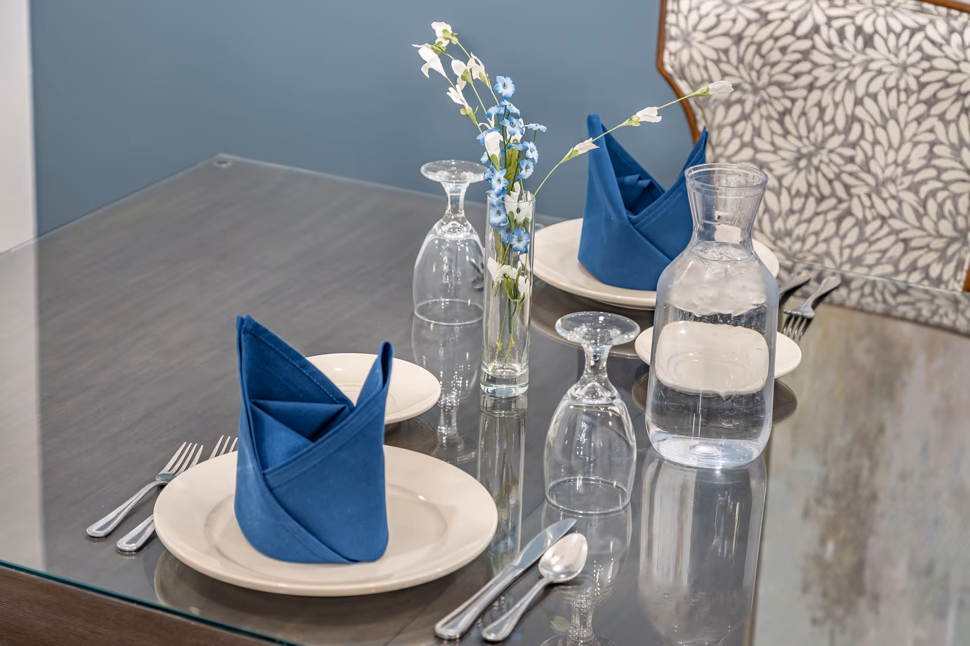 A dining table set for two with white plates, blue folded napkins, silverware, upside-down wine glasses, a glass carafe filled with water and ice, and a small vase with white and blue flowers. The table has a glass top and a chair with a floral patterned backrest is visible.