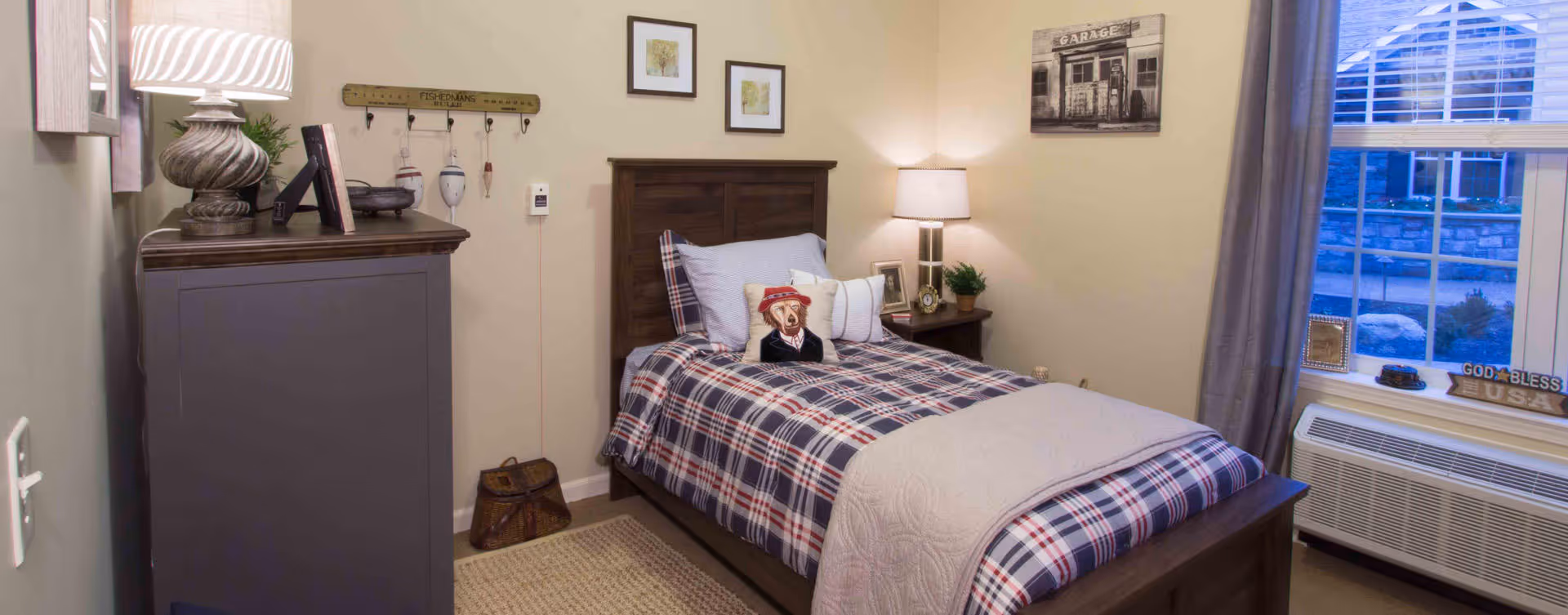 A cozy bedroom with a single bed featuring plaid bedding and a teddy bear wearing a hat and scarf. The room has a wooden dresser with a lamp and decorative items, a nightstand with a lamp and plant, framed pictures on the walls, a window with gray curtains, and an air conditioning unit below the window.