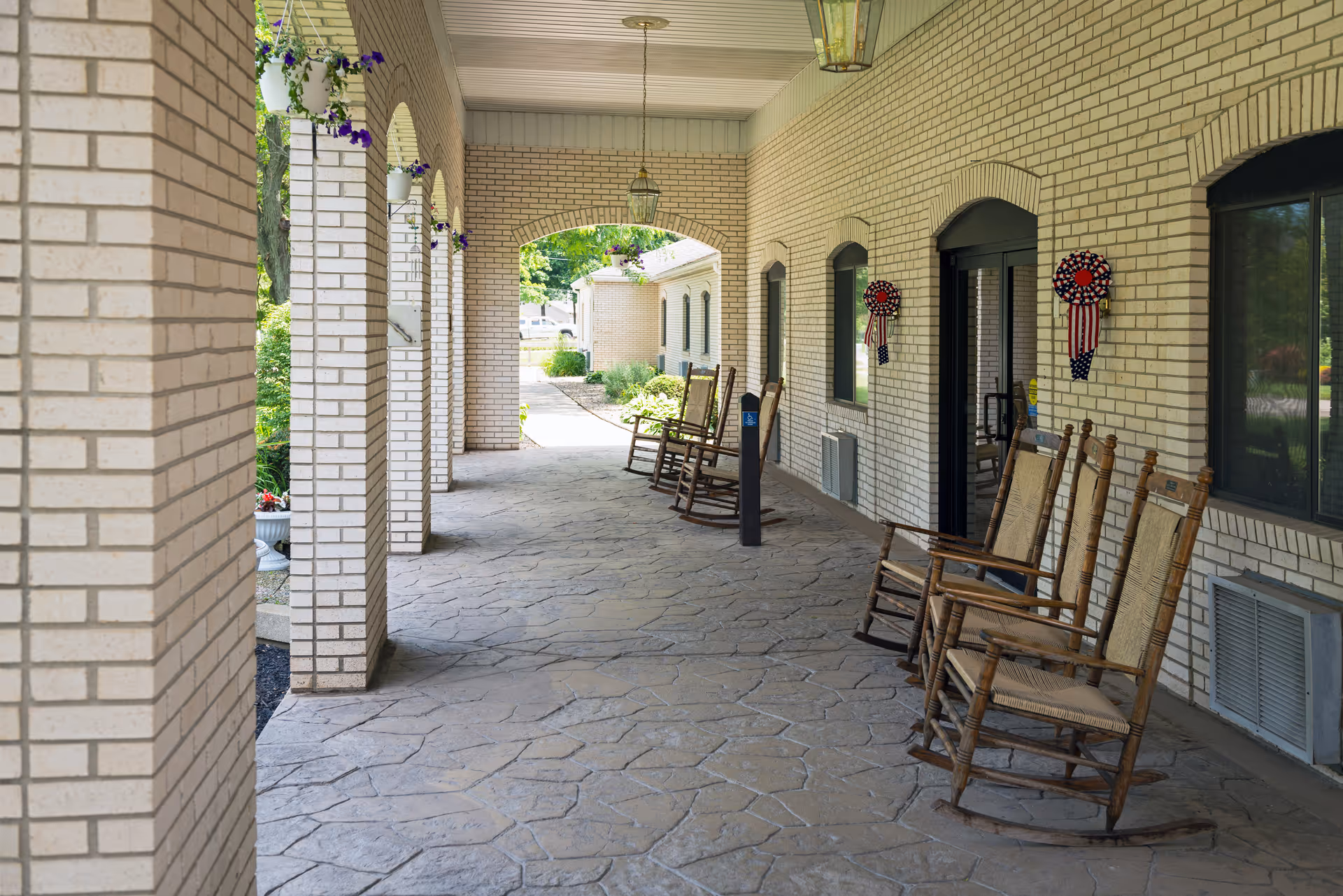 Covered outdoor walkway with beige brick walls and stone flooring, featuring several wooden rocking chairs lined up along the right side. Hanging flower pots with purple flowers are attached to the left side arches, and patriotic wreaths decorate the wall near the windows and door.