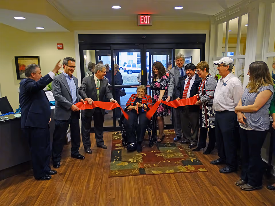 A group of people gathered inside a building for a ribbon-cutting ceremony. An elderly woman in a wheelchair is cutting a large red ribbon with the help of several men and women standing around her. The setting appears to be the entrance lobby of a facility with wooden floors, a patterned rug, and glass doors leading outside.