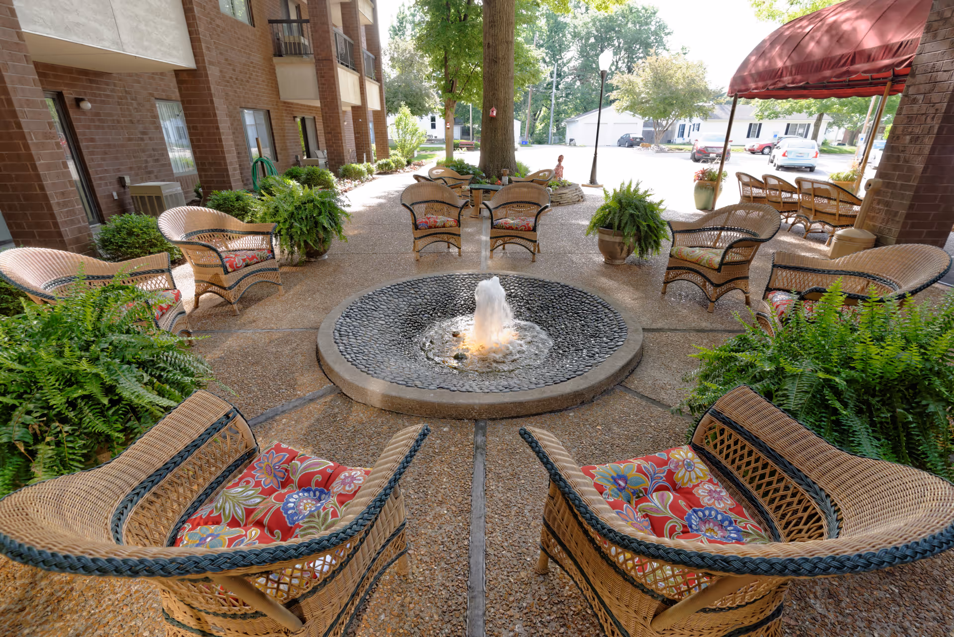 Outdoor seating area at Hickory Glen featuring wicker chairs with colorful floral cushions arranged around a circular stone fountain with water spouting in the center. The area is surrounded by potted ferns and greenery, with a brick building on the left and a red awning covering additional seating on the right. Trees and a parking area with cars are visible in the background.