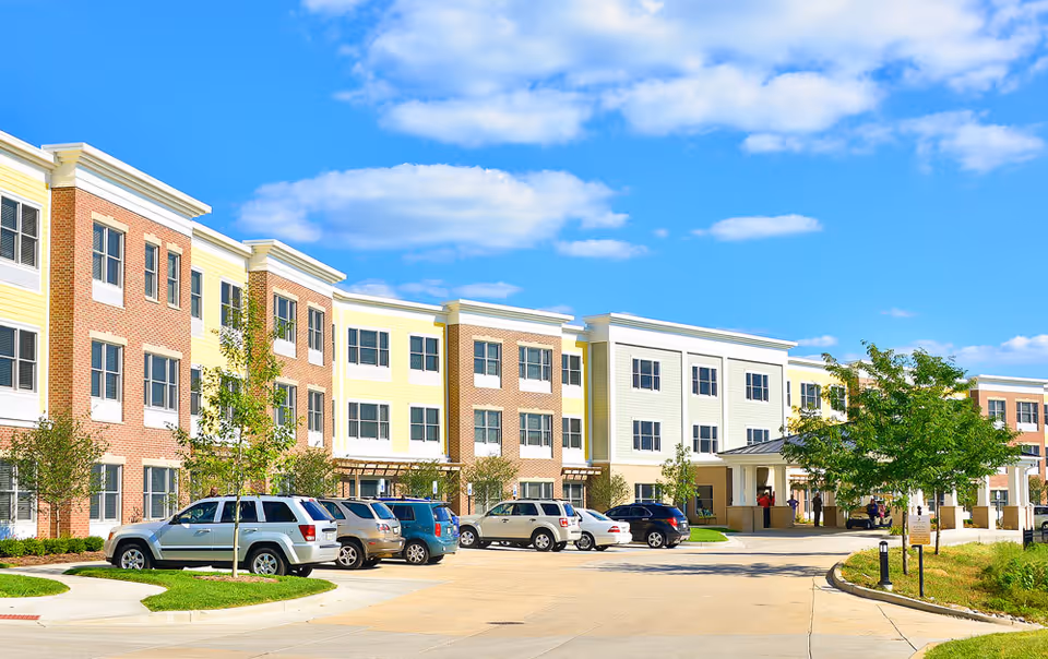 Exterior view of a multi-story senior living facility with a parking lot in front, several parked cars, and a few people near the entrance under a clear blue sky.