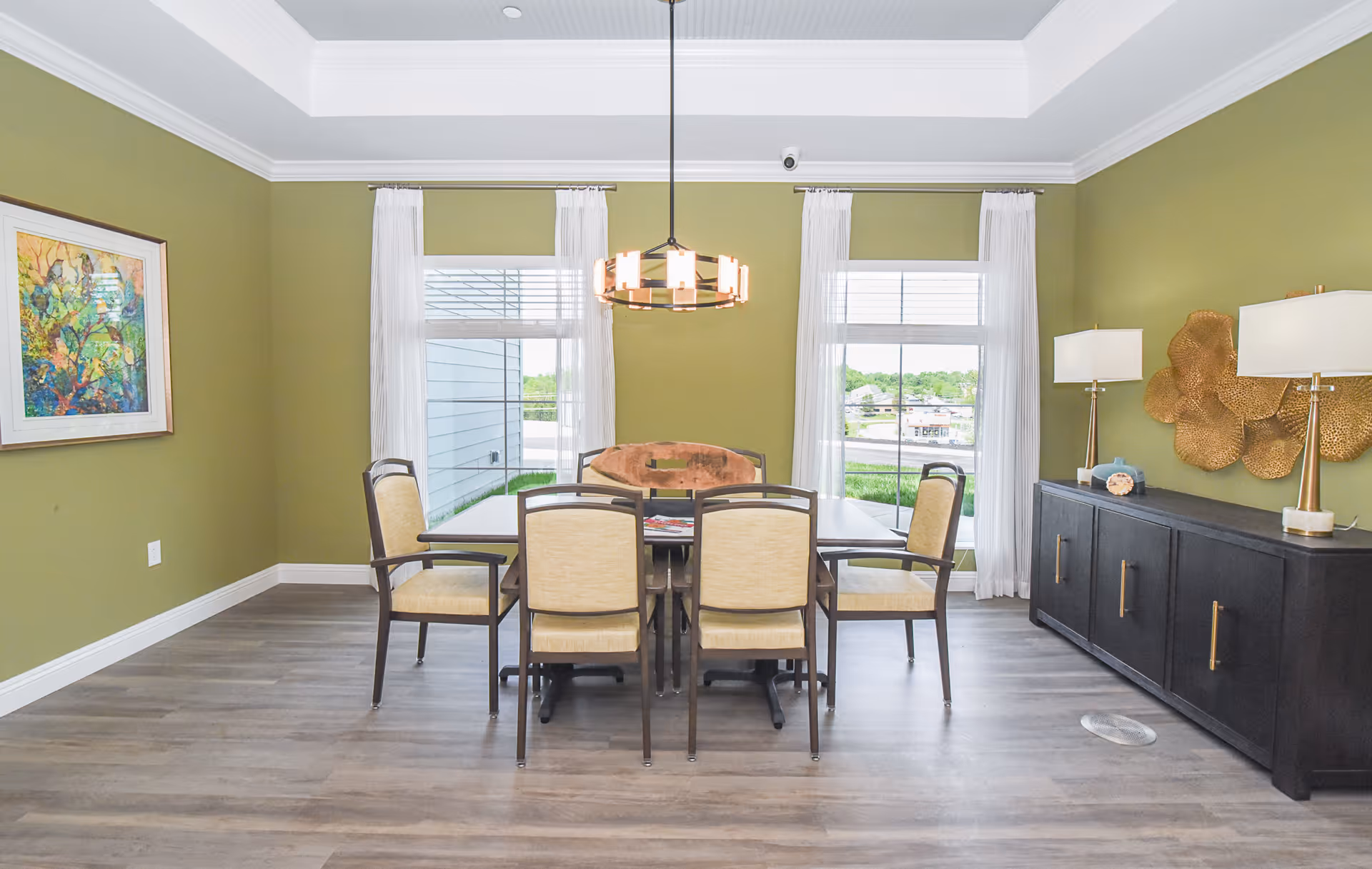 A dining room with green walls, two large windows with white curtains, a rectangular dining table with six beige cushioned chairs, a modern chandelier hanging above the table, a black sideboard with two lamps and decorative gold wall art, and a colorful framed painting on the left wall.