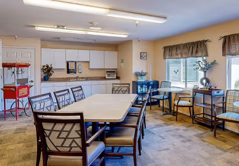 A dining area in a senior living facility with a long rectangular table surrounded by chairs in the center. In the background, there is a kitchenette with white cabinets, a countertop, a microwave, and a popcorn machine on the left side. To the right, there are smaller round tables with chairs near windows with brown valances, and decorative plants on a small cabinet and table.