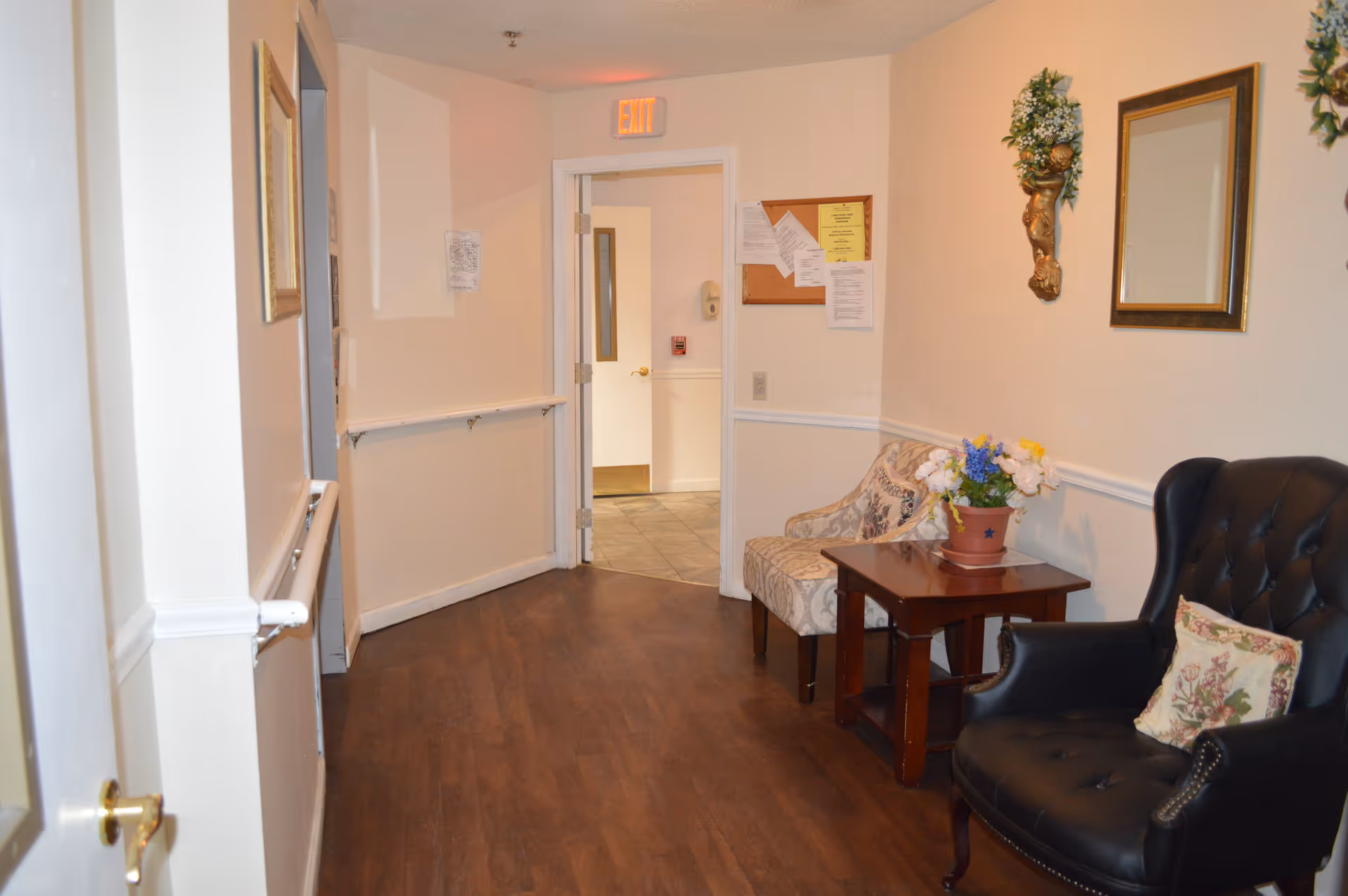 A hallway in a senior living facility with wooden flooring, beige walls, and white trim. There is a black leather armchair with a floral pillow, a patterned upholstered chair, and a wooden side table with a potted flower arrangement. A mirror and decorative wall sconces with greenery are mounted on the wall. At the end of the hallway, there is an open door leading to a tiled area with an exit sign above the door.