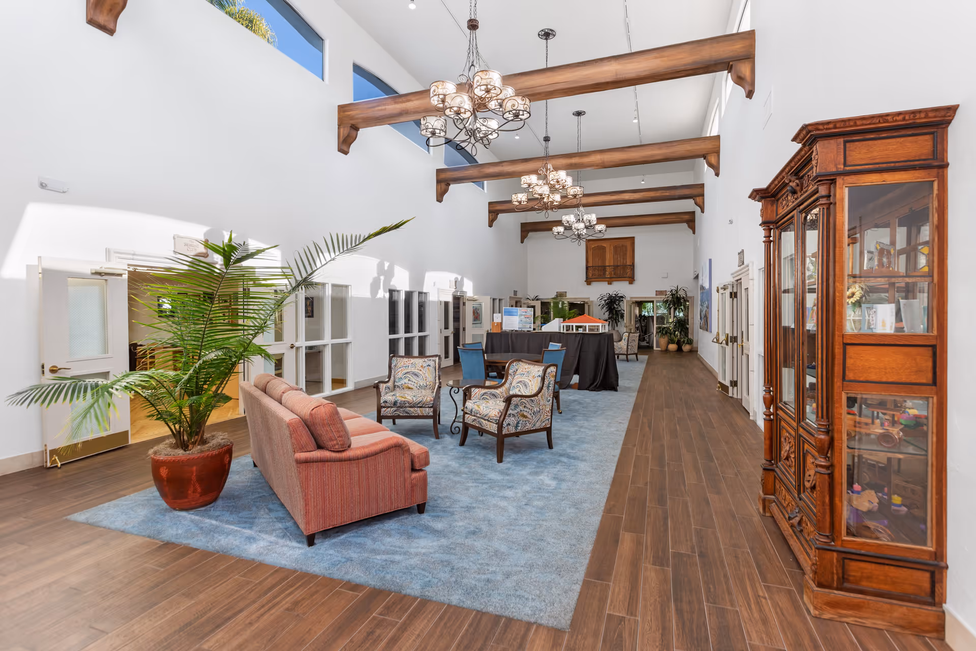 A spacious, well-lit interior lounge area with high ceilings and wooden beams. The room features multiple chandeliers, a red sofa, patterned armchairs, a large potted plant, and a wooden display cabinet on the right. The floor is a combination of wood and a blue carpet, and there are several doors and windows along the walls.