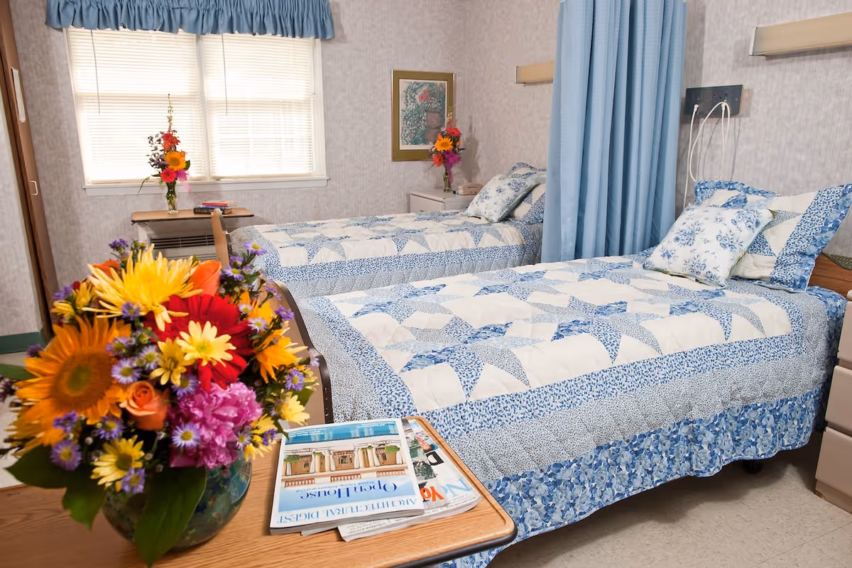 Two twin beds with blue floral patchwork quilts in a bright nursing-home bedroom, a bouquet of colorful flowers and magazines on a bedside table.
