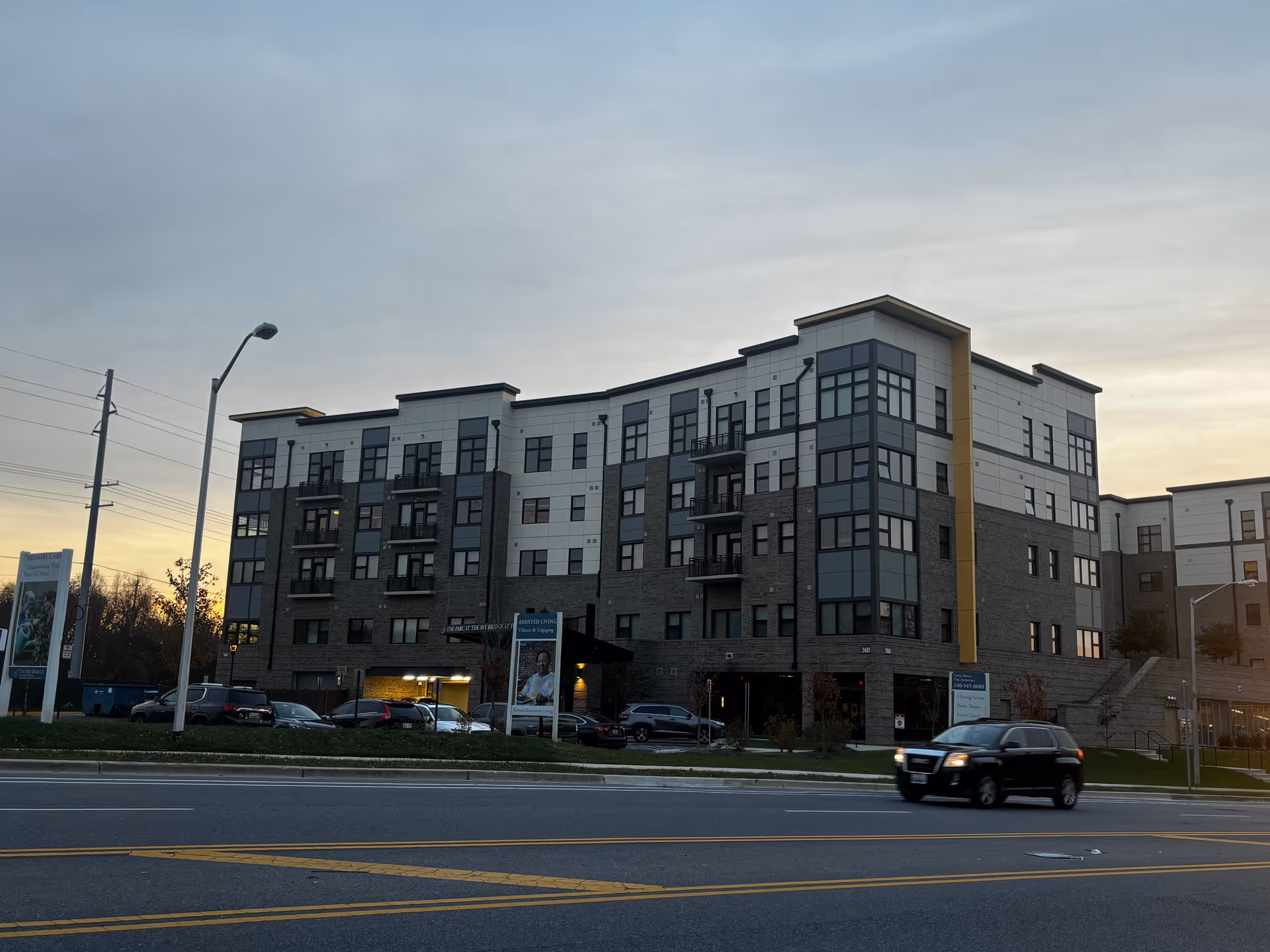 Exterior view of a modern multi-story senior living facility named Sky Bridge at Town Center during dusk, with several cars parked in front and a black SUV driving on the road in the foreground.