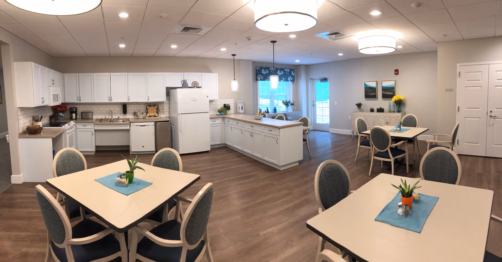 A bright and clean communal kitchen and dining area with white cabinetry, a refrigerator, dishwasher, and microwave. There are several tables with chairs arranged around the room, each table decorated with a small plant and a blue placemat. The room has wood flooring, recessed lighting, pendant lights, and a window with a blue floral valance. A door leads outside or to another room, and there are framed pictures and a cabinet with flowers on the far wall.