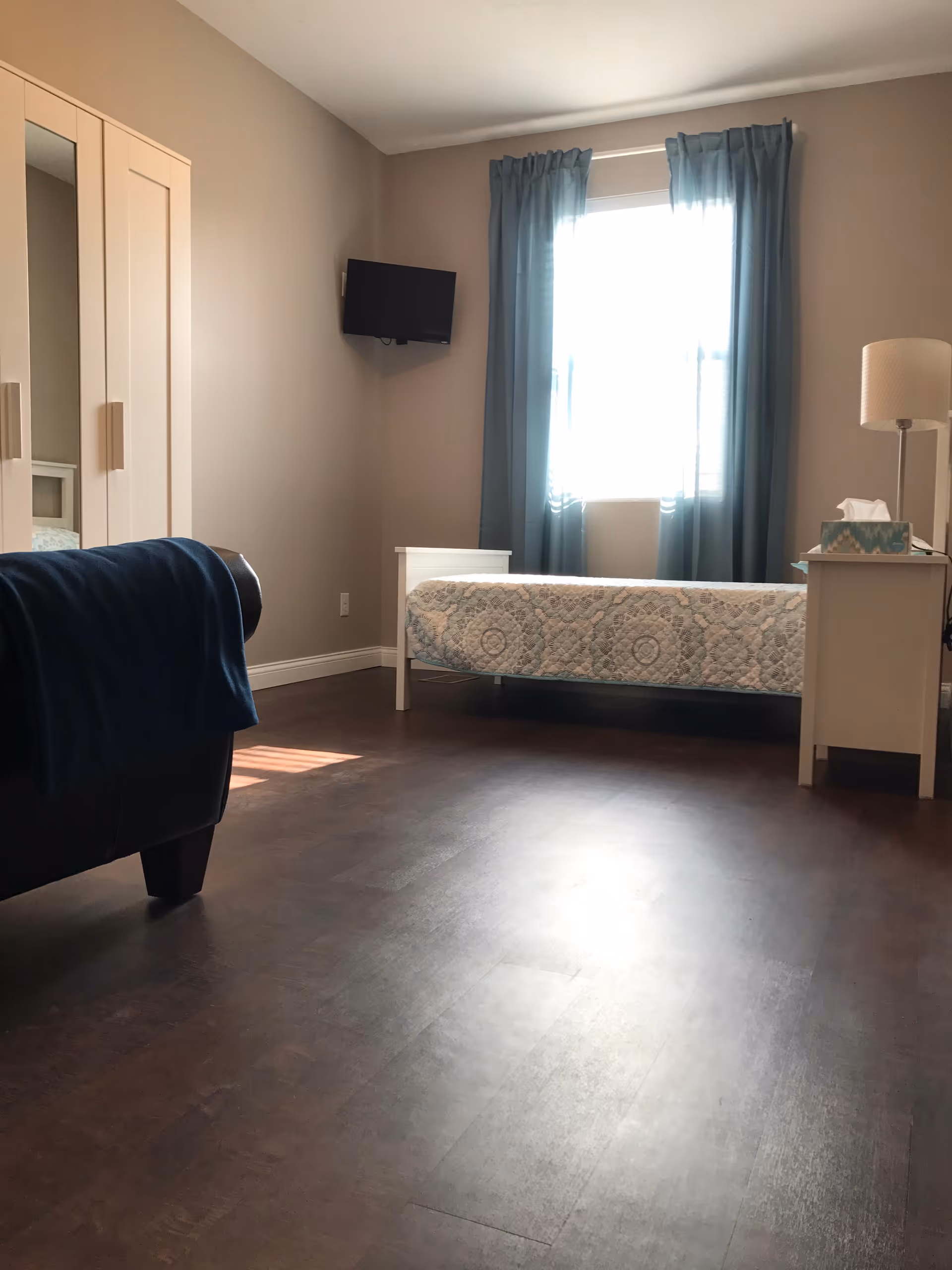 A bright bedroom with a single bed covered in a patterned quilt, a nightstand with a lamp and tissue box, a wall-mounted TV, a wardrobe with mirrored doors, and a window with blue curtains letting in natural light.
