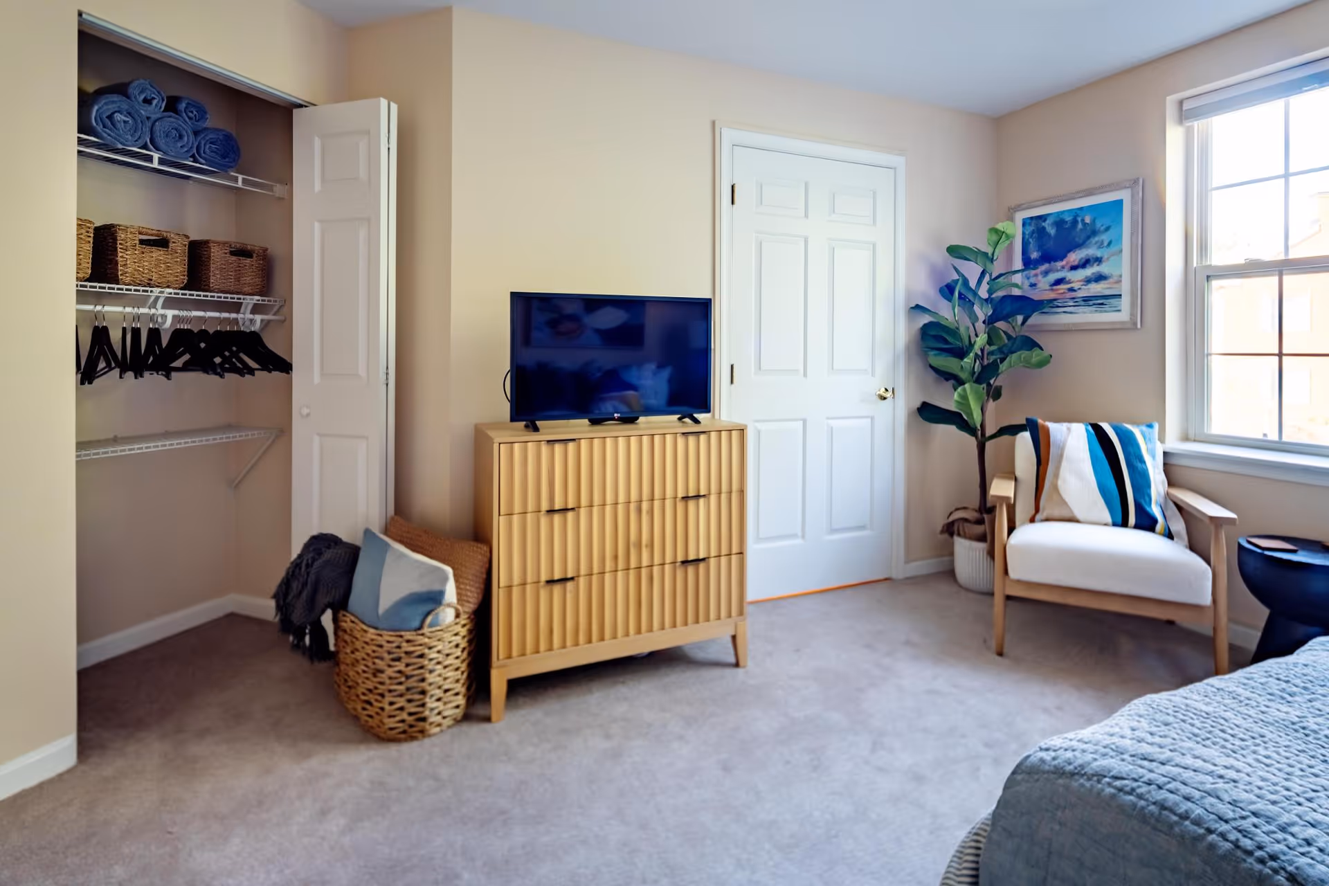 A cozy bedroom featuring a wooden dresser with a flat-screen TV on top, an open closet with shelves holding rolled blue towels and wicker baskets, and black hangers on a rod. A woven basket with blankets is placed on the floor next to the dresser. There is a comfortable armchair with a striped pillow near a window that lets in natural light. A potted plant and framed artwork decorate the corner of the room.