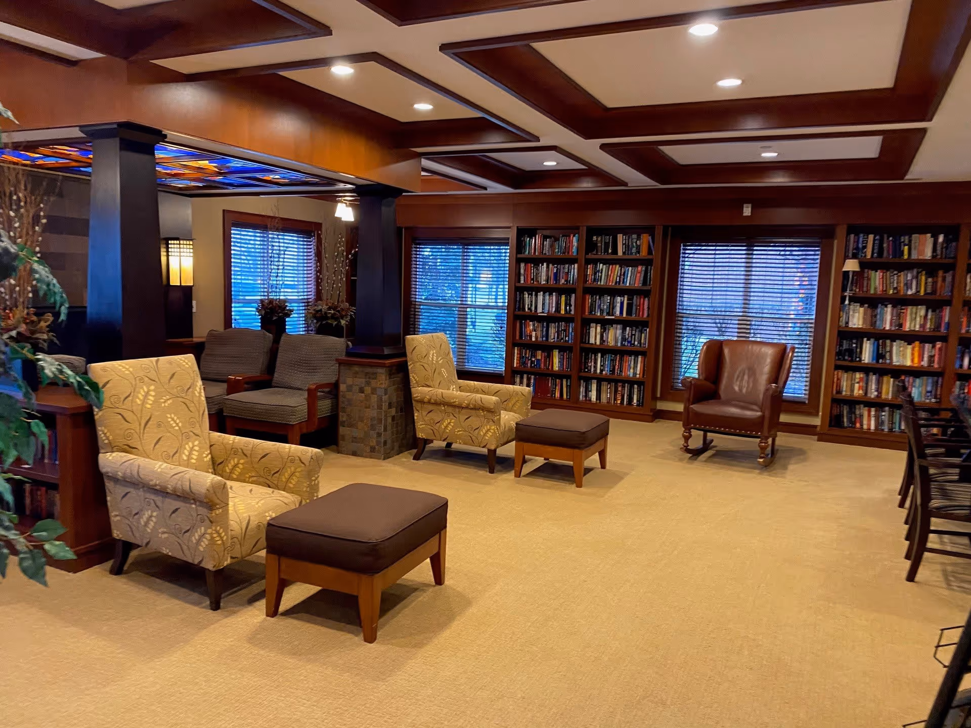 A cozy library or reading room with beige carpet, two patterned armchairs with matching ottomans, a brown leather armchair, and several bookshelves filled with books. The room features wooden beams on the ceiling, large windows with blinds, and soft lighting.