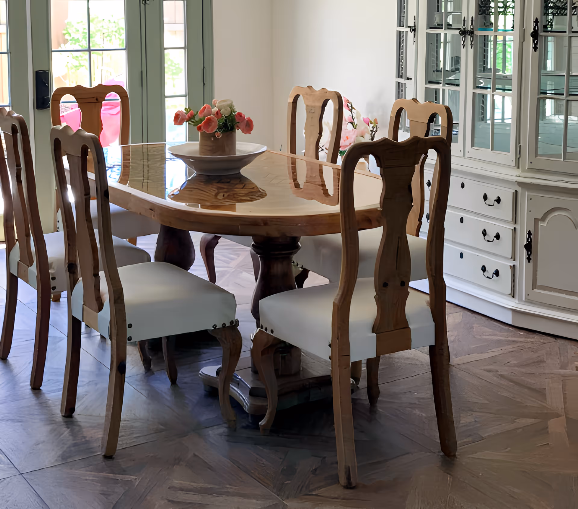 A dining room with a wooden dining table and six matching wooden chairs with white cushions. A vase with pink and white flowers is placed on the table. In the background, there is a white cabinet with glass doors and drawers, and a door with glass panels letting in natural light.
