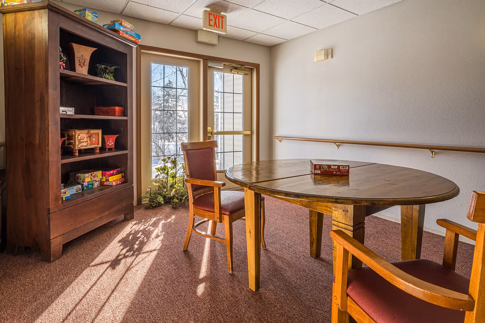 A small room with a round wooden table and three wooden chairs with red cushions. A wooden bookshelf filled with board games and decorative items is against the left wall. Sunlight streams through glass doors with an exit sign above them, casting shadows on the carpeted floor. A handrail runs along the right wall.