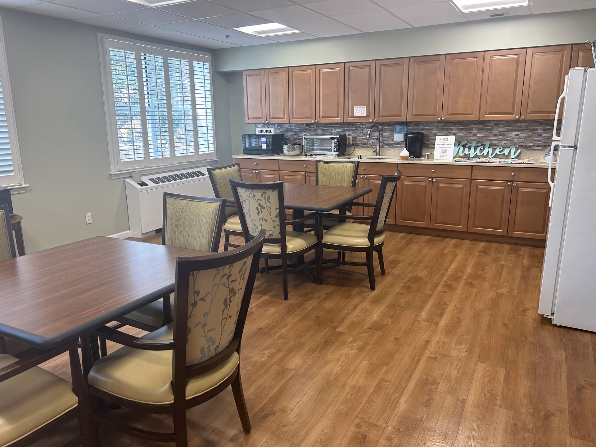 Communal dining area with wooden tables and chairs and a kitchenette with cabinets and appliances.