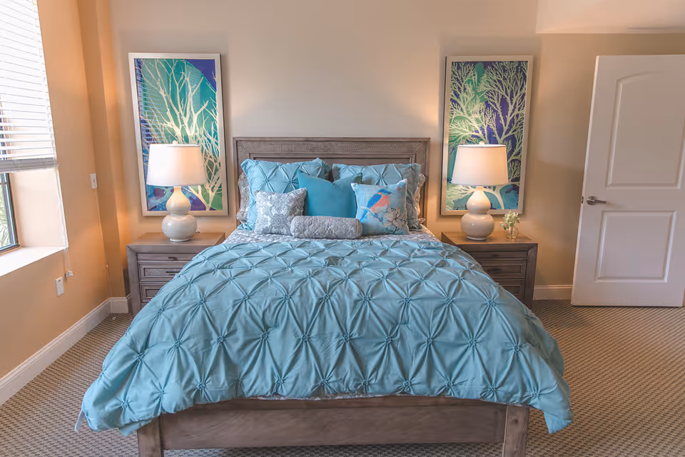 A neatly made bed with a light blue comforter and multiple decorative pillows in a bedroom. The bed has a wooden headboard and is flanked by two wooden nightstands, each with a white lamp. Above each nightstand hangs a framed artwork featuring tree designs in blue and green tones. A window with blinds is on the left side of the room, and a white door is partially open on the right side.