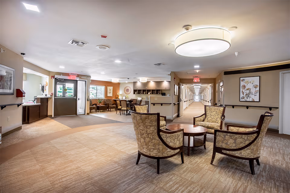 A spacious and well-lit common area in a senior living facility with four upholstered armchairs arranged around a circular wooden coffee table. The room features beige walls, carpeted flooring with a circular pattern, framed floral artwork, and a long hallway leading to other rooms. There is a reception desk area near the entrance with large windows allowing natural light to enter.