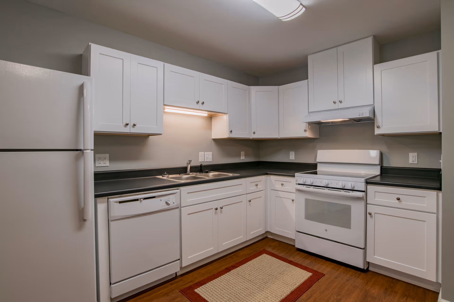 A modern kitchen with white cabinets, a white refrigerator, a white dishwasher, a white stove with an oven, and a stainless steel double sink. The countertops are dark, and there is a small rectangular rug on the wooden floor. The walls are painted gray, and there is a ceiling light fixture.