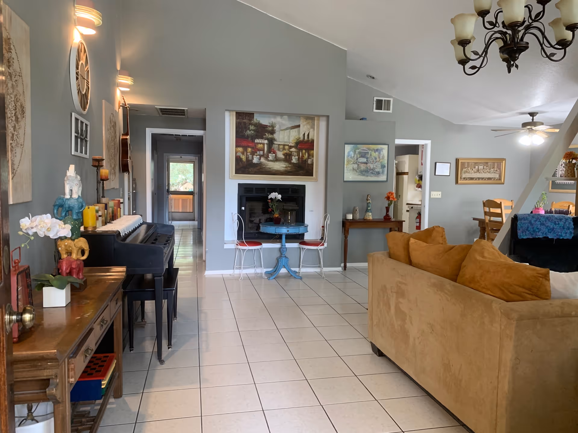A spacious living room with beige tiled floors and light gray walls. The room features a tan sofa with brown cushions, a black piano with a bench, a small blue round table with two white chairs in front of a fireplace, and various wall art and decorations. There is a wooden dining table with chairs visible in the background, along with ceiling fans and a chandelier lighting fixture.