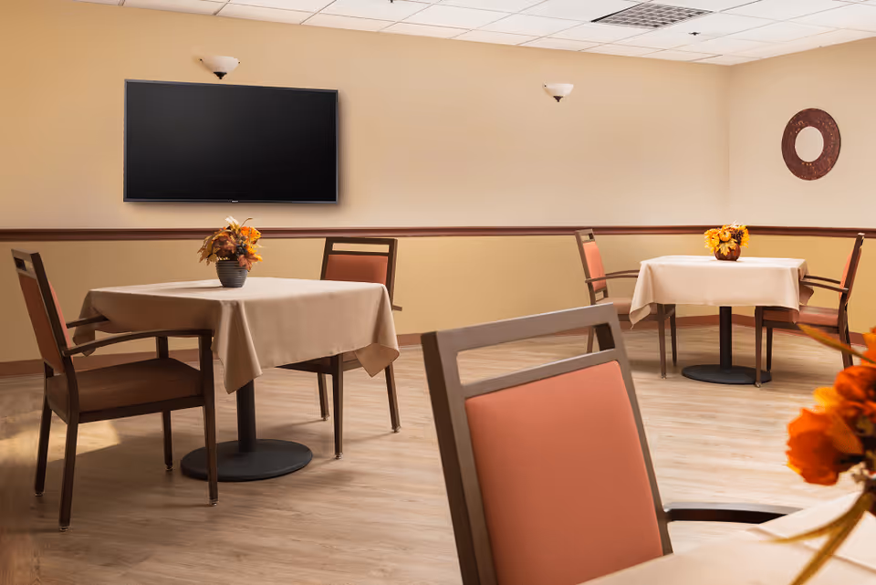A small dining area with three tables covered with beige tablecloths, each table having a floral centerpiece. The room has light-colored walls with a darker trim, a wall-mounted flat screen TV, and a circular decorative wall piece. The floor is wood, and the chairs have wooden frames with orange cushions.