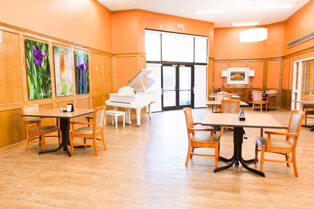 A spacious, well-lit common area with wooden floors and orange walls featuring wood paneling. The room contains several tables with chairs arranged around them, a white grand piano with a matching bench, and colorful floral artwork on the walls. Large windows and glass doors allow natural light to enter the room.