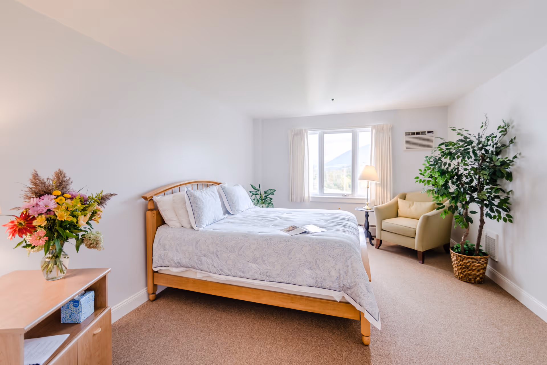 A bright bedroom with a wooden bed frame and white patterned bedding, a small wooden nightstand with a colorful flower arrangement, a beige armchair next to a window with cream curtains, a floor lamp, and a large potted plant in the corner.
