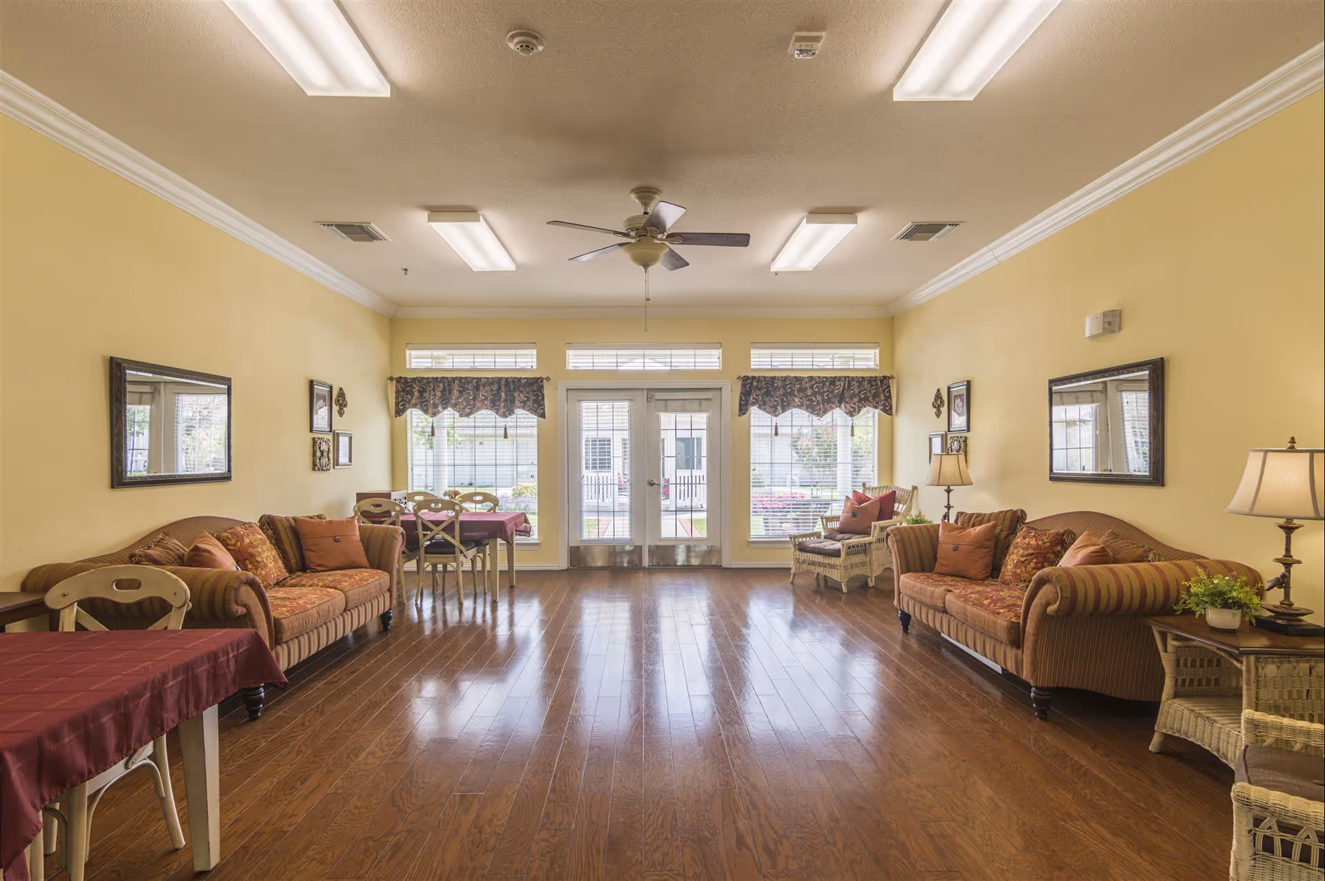 A bright and spacious living room with yellow walls and wooden flooring. The room features two patterned sofas with cushions, a wicker chair with a cushion, two tables with burgundy tablecloths, and several chairs. There are two large mirrors on the walls, framed pictures, table lamps, and a ceiling fan. Large windows and a glass door at the far end let in natural light and offer a view of an outdoor area.
