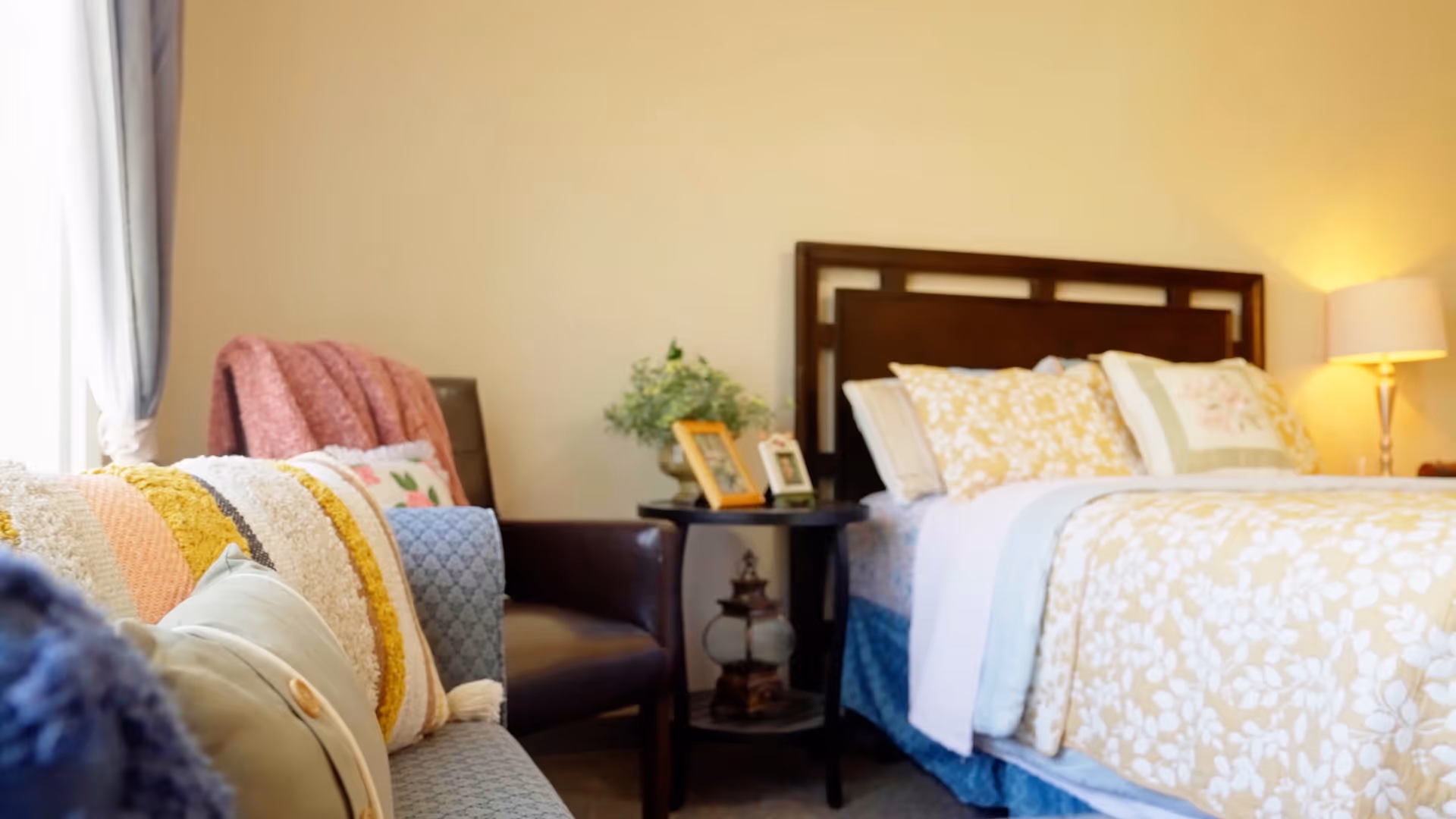 A cozy bedroom featuring a bed with floral patterned bedding and multiple pillows, a bedside table with framed photos and a small plant, a lamp providing warm light, and a comfortable armchair with a pink throw blanket near a window with curtains.