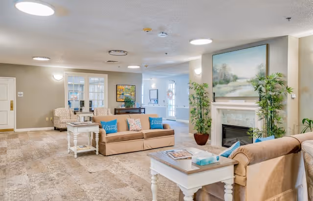 A spacious and well-lit living room area in a senior living facility featuring two beige sofas with blue and floral throw pillows, a white coffee table with magazines and a tissue box, a fireplace with a large landscape painting above it, and potted plants on either side of the fireplace. The room has beige carpeting, neutral-colored walls, and additional seating with two patterned armchairs near a windowed wall.
