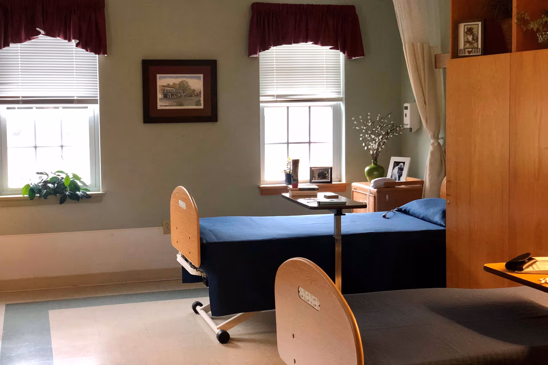Patient room with two hospital-style beds, windows with valances, a bedside table and cabinetry with decorative items.