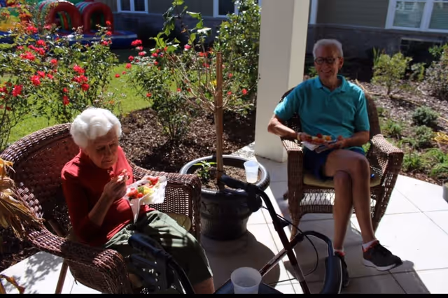 Two elderly individuals sitting outdoors on wicker chairs under a covered patio, enjoying a meal. One person is wearing a red top and green pants, and the other is wearing a teal shirt and shorts. There is a walker and a potted plant between them, with a garden and building visible in the background.