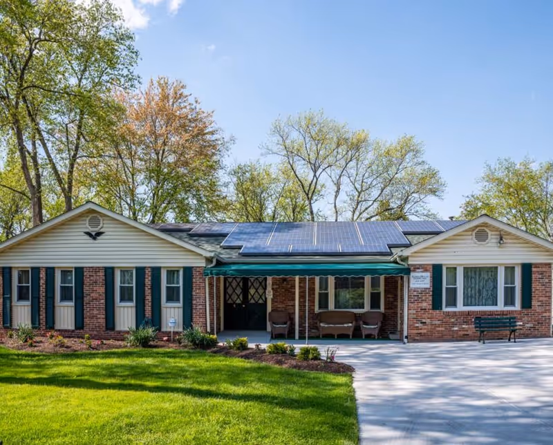 Front exterior of a single-story brick senior living building with solar panels on the roof, a covered entrance with seating, and a green lawn.