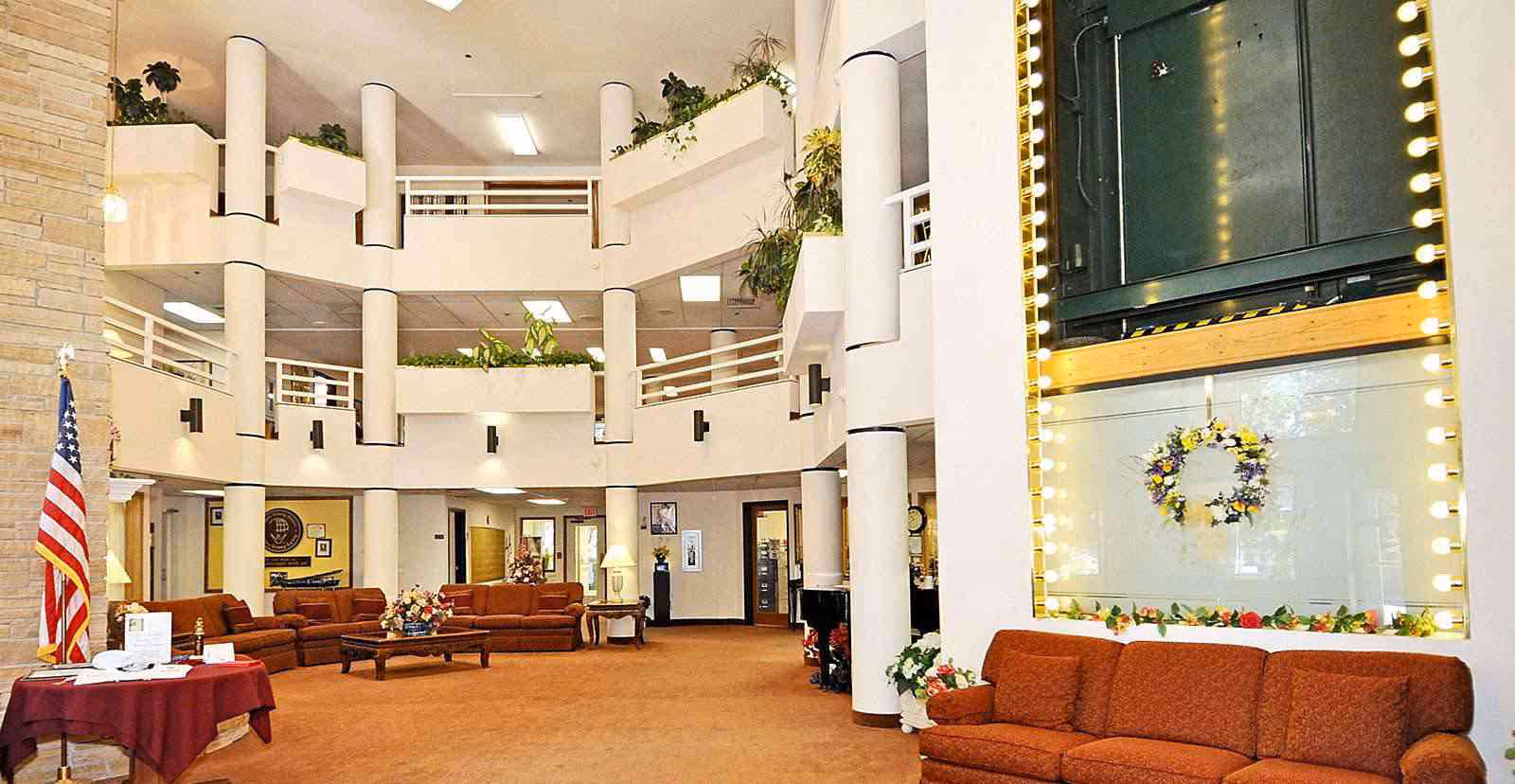 Spacious multi-level senior living facility atrium with sofas, columns, plants, and an American flag.