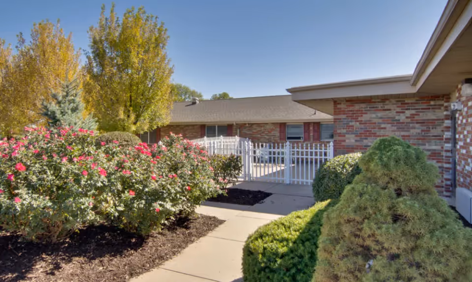 Outdoor view of a senior living facility courtyard with a paved walkway, well-maintained bushes, flowering plants with pink blooms, and trees with autumn foliage. The building has a brick exterior with red shutters and a white picket fence enclosing a patio area.