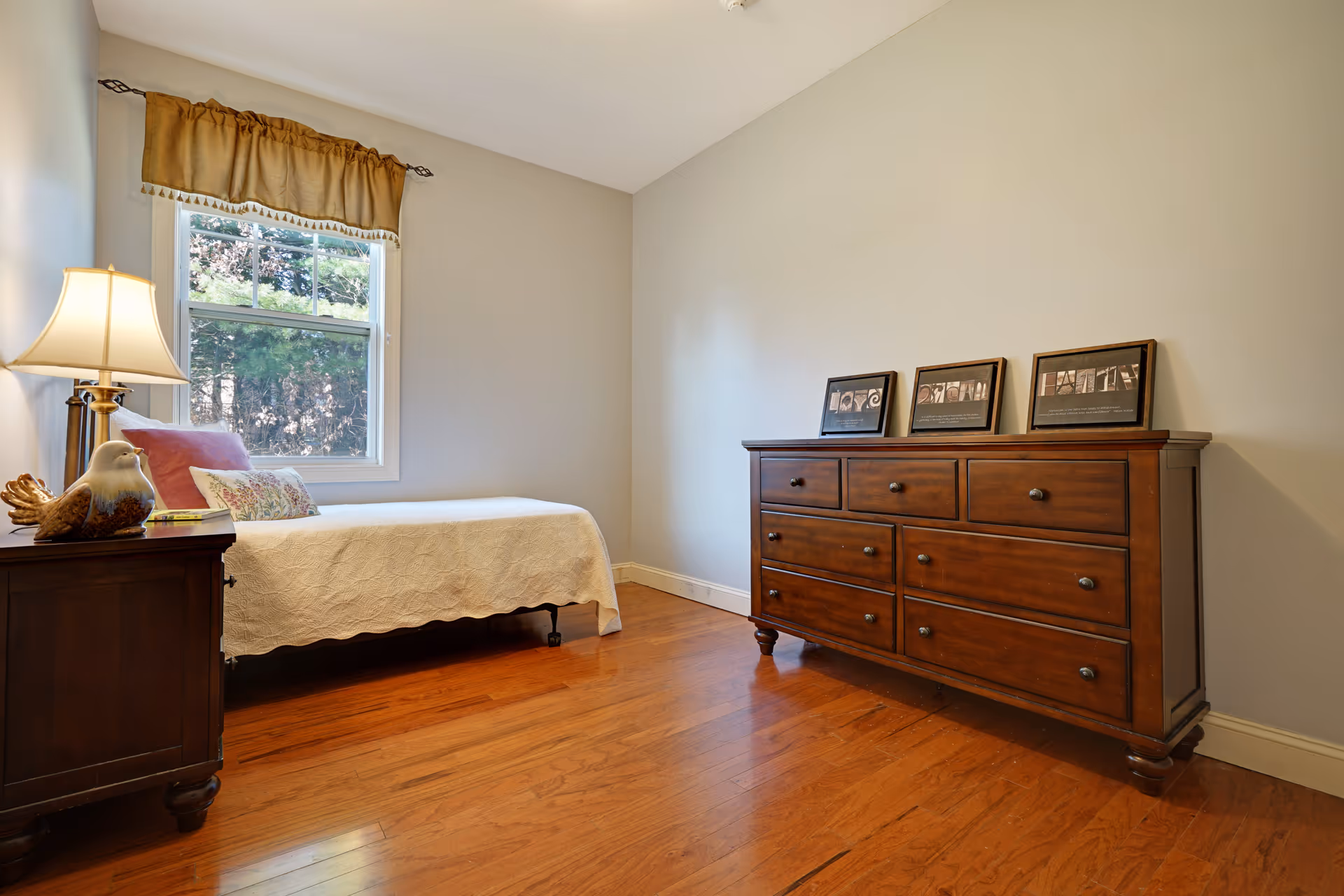 A simple bedroom with a single bed covered in a white quilt, a wooden nightstand with a lamp and decorative bird figurine, a window with a brown valance, and a large wooden dresser with framed pictures on top. The room has wooden flooring and light-colored walls.