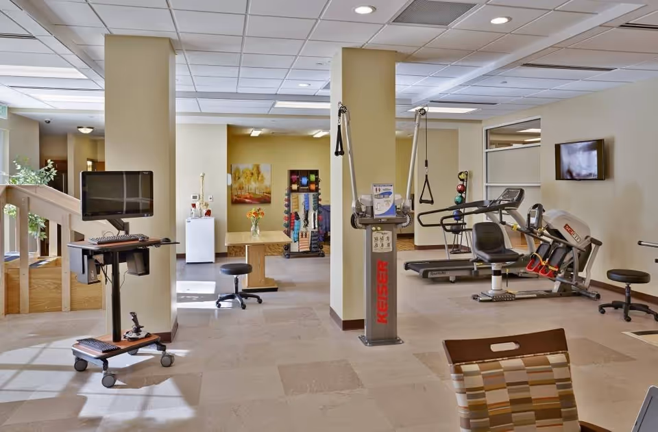 A well-lit fitness and rehabilitation room in a senior living facility featuring exercise equipment including a Keiser machine, a treadmill, and a recumbent bike. There is a computer workstation on wheels to the left, a table with a stool in the background, and a rack with colorful exercise bands and weights. The room has beige walls, a tiled floor, and a ceiling with recessed lighting.